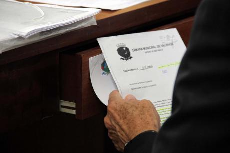 #PraCegoVer: Foto mostra vereador segurando um documento, sentado em sua cadeira.