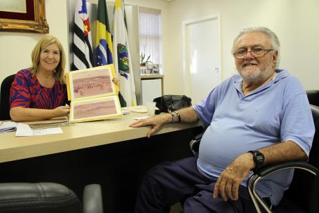 #PraCegoVer: Ex-prefeito Arildo e presidente da Câmara, Dalva Berto estão sentados à mesa da presidência sorrindo para a foto. Ela segura um álbum de fotografias em preto e branco. Ao fundo, estão bandeiras coloridas.