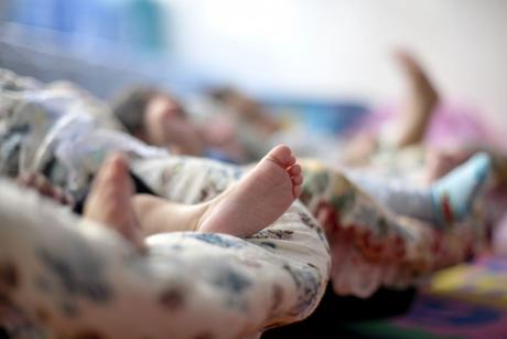 #PraCegoVer: Foto mostra pernas de neném deitado em um bebê-conforto. Ao fundo, desfocados, vêem-se outros bebês-conforto com crianças deitadas dentro. A imagem parece ter sido tirada em um abrigo ou creche para recém-nascidos.