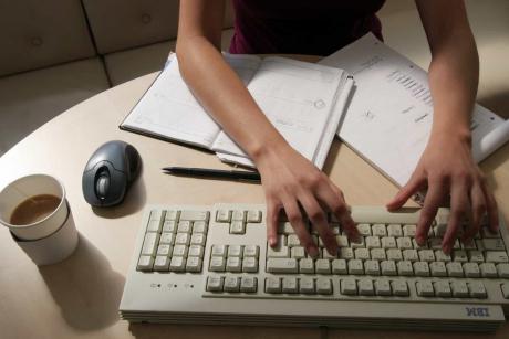 #PraCegoVer: Foto mostra braços e mãos de pessoa usando o teclado de um computador, junto com folhas de papel e caderno de trabalho, ao lado de um copo de papel com café com leite.