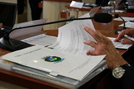 #PraCegoVer: Foto mostra as mãos de um vereador folheando documentos durante a sessão.