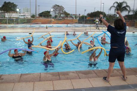 #PraCegoVer: Foto mostra educadora física levantando os braços e dando as instruções para uma turma de praticantes de hidroginástica em uma piscina. A turma veste trajes de banho e usa boias de espuma.