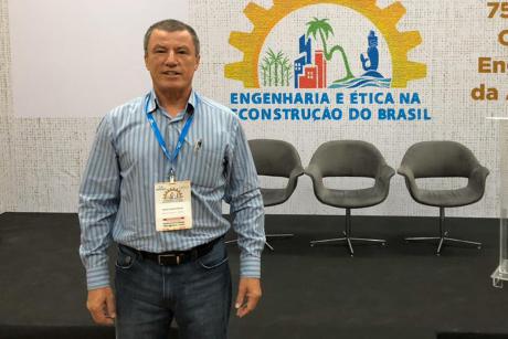 #PraCegoVer: Mário Antonio Masteguin, vestido com camisa e calça jeans e usando um crachá típico de conferências, posa para foto em frente a painel onde se lê "Engenharia e Ética na Reconstrução do Brasil"