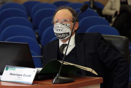 #PraCegoVer: Vereador Conti acompanha a sessão de sua tribuna na Câmara. Ele usa máscara e tem uma pasta aberta à sua frente. Ao fundo, veem-se as cadeiras do plenário vazias. 