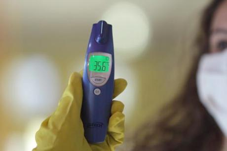 #PraCegoVer: Foto mostra a mão de uma pessoa com luva amarela segurando um termômetro azul, que marca 35,6 graus. No desfoque, ao fundo, há parte do rosto de uma pessoa com máscara.