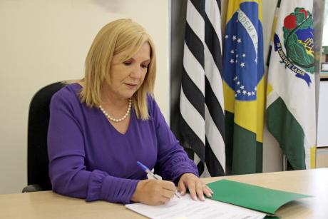#PraCegoVer: Dalva Berto assina um papel fixado de uma pasta verde. Ela senta-se a uma mesa. Ao fundo, estão bandeiras do estado de São Paulo, do Brasil e de Valinhos.