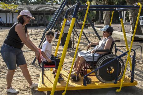 #PraCegoVer: Em um parque, uma mulher empurra um balanço adaptado para crianças com deficiência. Sobre o balanço, há um menino que parece ter Síndrome de Down e uma menina cadeirante. O equipamento é uma plataforma metálica fixada a uma estrutura tubular. 