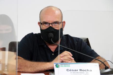 #PraCegoVer: César Rocha fala ao microfone em seu assento na mesa diretora da Câmara. Ele usa uma máscara negra. Em frente dele há uma placa com o seu nome 