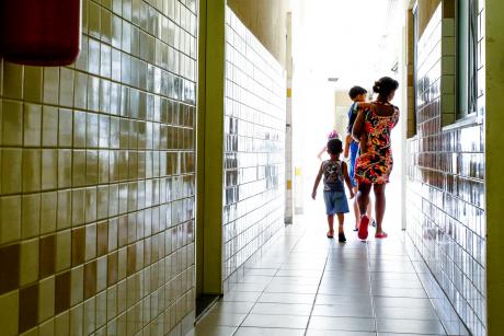 #PraCegoVer: Mulher caminha em direção à luz em corredor revestido com azulejos. Ela carrega um menino no colo e está acompanhada de mais três crianças. Foto de uma casa-abrigo para mulheres vítimas de violência em Belford Roxo (RJ). 