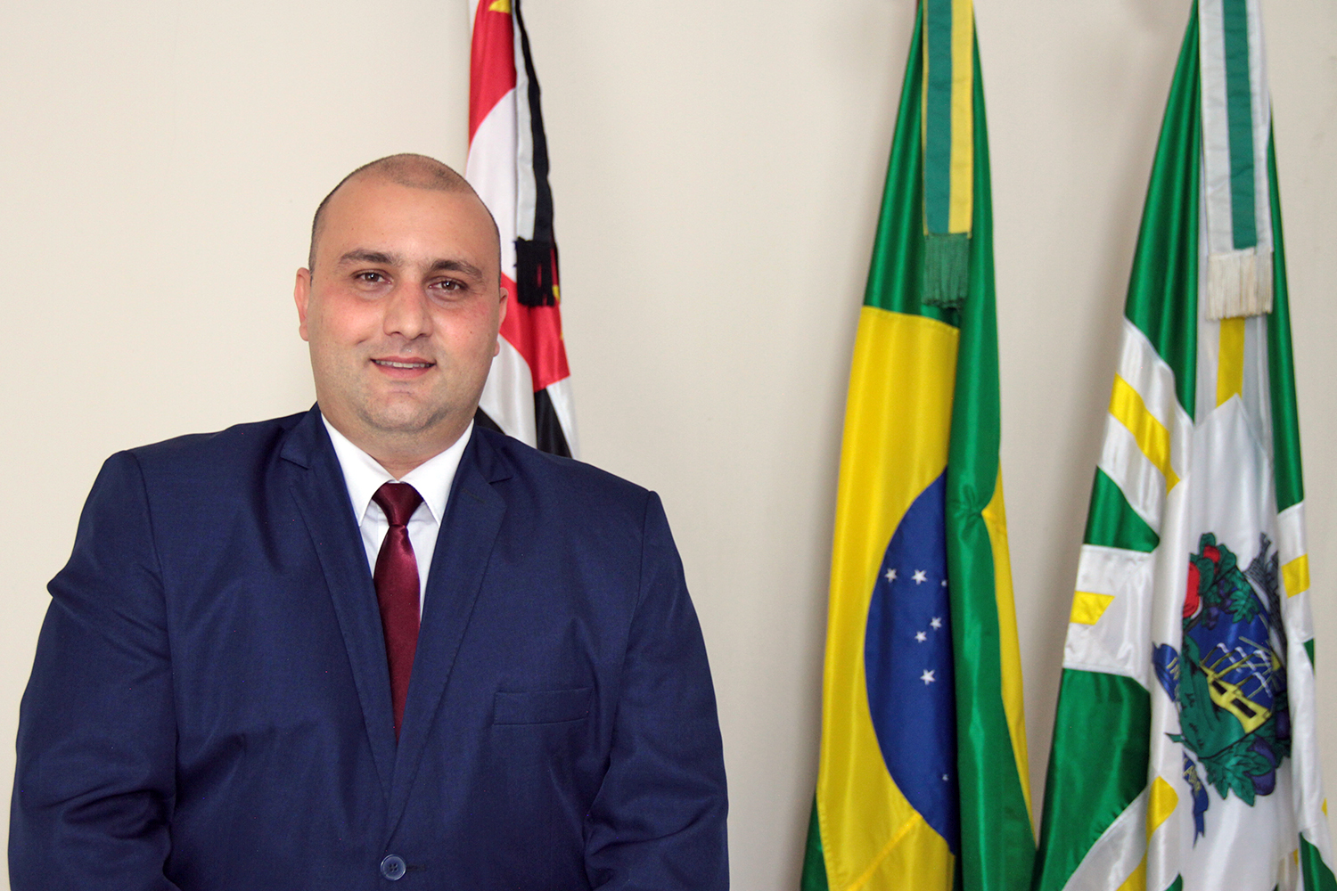 #PraCegoVer: Vereador Thiago Samasso posa e sorri para foto em frente às bandeiras de São Paulo, Brasil e Valinhos.