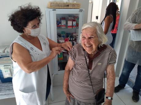 #PraCegoVer: Foto mostra profissional de saúde dando injeção em uma idosa sorridente. Ao fundo, há um armário para armazenagem de medicamentos. 