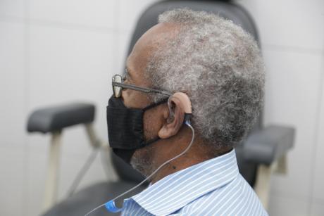 #PraCegoVer: Homem de cabelos brancos usa aparelho auditivo em seu ouvido esquerdo.