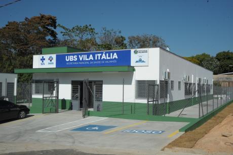 #PraCegoVer: UBS da Vila Itália, um edifício de um andar, cercado por grades e pintado de branco e verde. Uma placa azul traz o nome da UBS, junto com diversas logomarcas governamentais. 