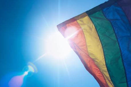 #PraCegoVer: Bandeira do arco-íris, símbolo do movimento LGBTQIA+, é agitada contra céu azul e sol claro. 