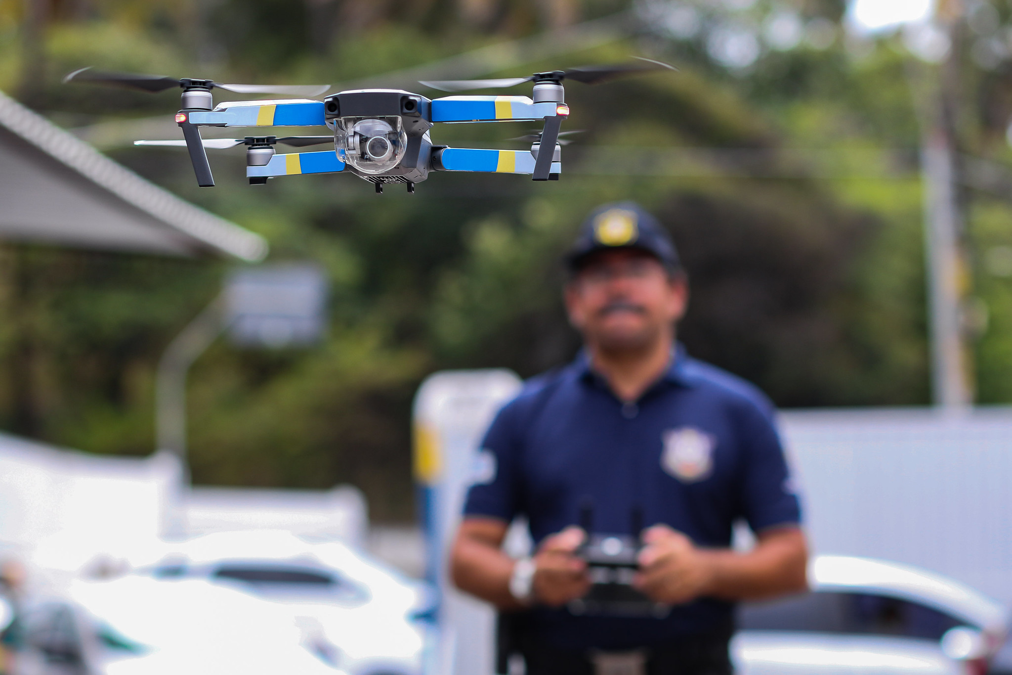 #PraCegoVer: Enxerga-se em primeiro plano um drone, aparelho com quatro hélices e câmera. Ao fundo, desfocado, vê-se um guarda civil municipal pilotando-o com o auxílio de um controle com antenas.