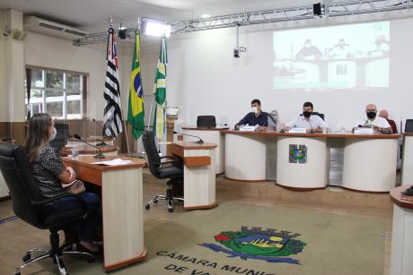 #PraCegoVer: Foto mostra Vânia Peretti sentada no plenário da Câmara, ouvindo as perguntas do vereador Gabriel Bueno. Ao lado de Gabriel, estão os vereadores André Amaral à direita e César Rocha à esquerda.