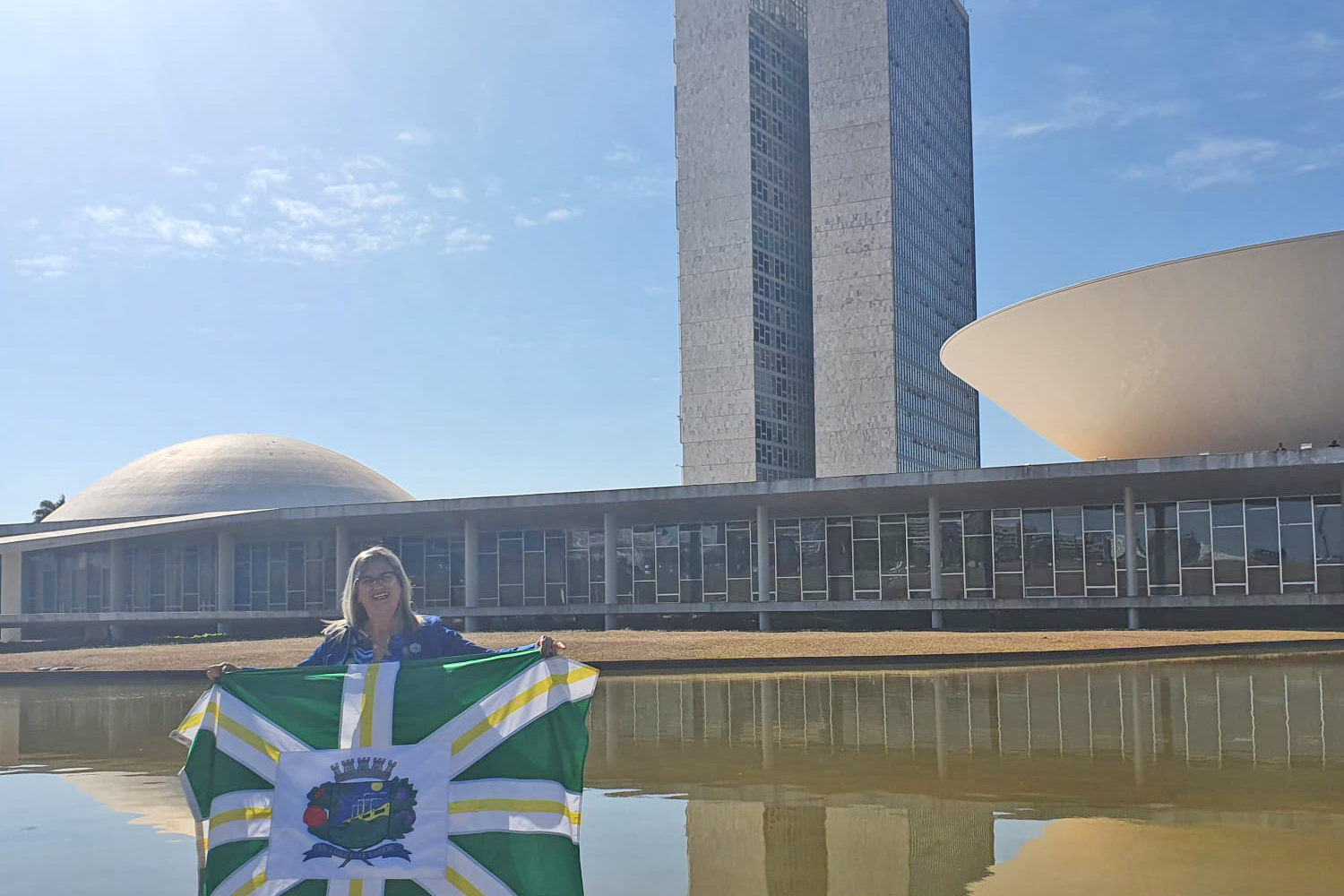 #PraCegoVer: Foto mostra a vereadora Simone Bellini em frente ao Congresso Nacional, segurando a bandeira de Valinhos.