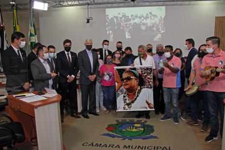 #PraCegoVer: Foto mostra vereadores e integrantes do Samba da Tia Rê posando para foto. No centro há um banner com foto de Regina Jesuíno, que deu nome ao grupo.