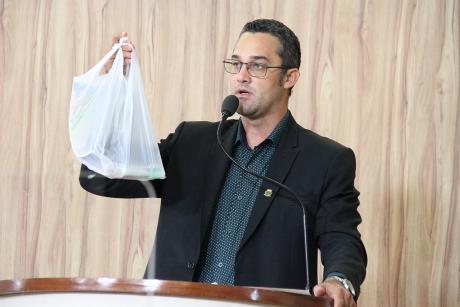 #PraCegoVer: Foto mostra o vereador Alécio Cau discursando na tribuna da Câmara. Ele mostra uma sacola plástica com alguns itens de material escolar dentro.