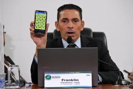 #PraCegoVer: Foto mostra o presidente da Câmara, vereador Franklin, segurando um celular que mostra o aplicativo da Câmara em funcionamento. Ele está em seu lugar no plenário, durante a sessão ordinária.