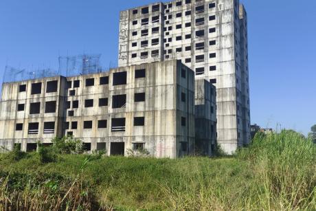 #PraCegoVer: Foto mostra dois prédios abandonados. A construção está com a estrutura evidente, sem pintura e sem janelas. Em volta, há mato alto.