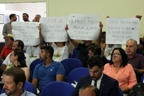 #PraCegoVer: foto mostra moradores do bairro Alpinas acompanhando a sessão. Eles erguem quatro cartazes que pedem solução para problemas diversos do bairro.