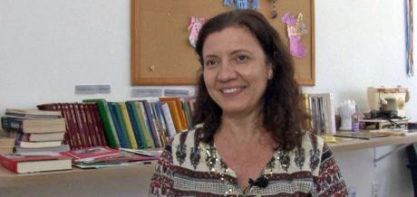 #PraCegoVer: Silene Gianechini sorri, sentada em frente a livros e a utensílios para atividades manuais no Creaps